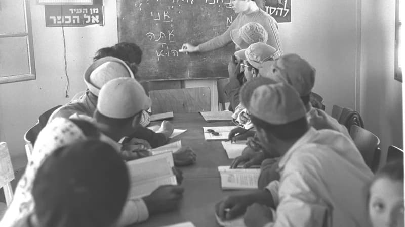 Language class students studying at a table while a teacher writes on the blackboard — comparing futur proche and futur simple in French